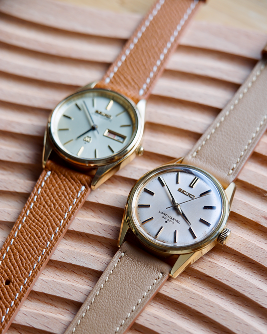 Two watches with brown and beige leather straps on a wooden surface
