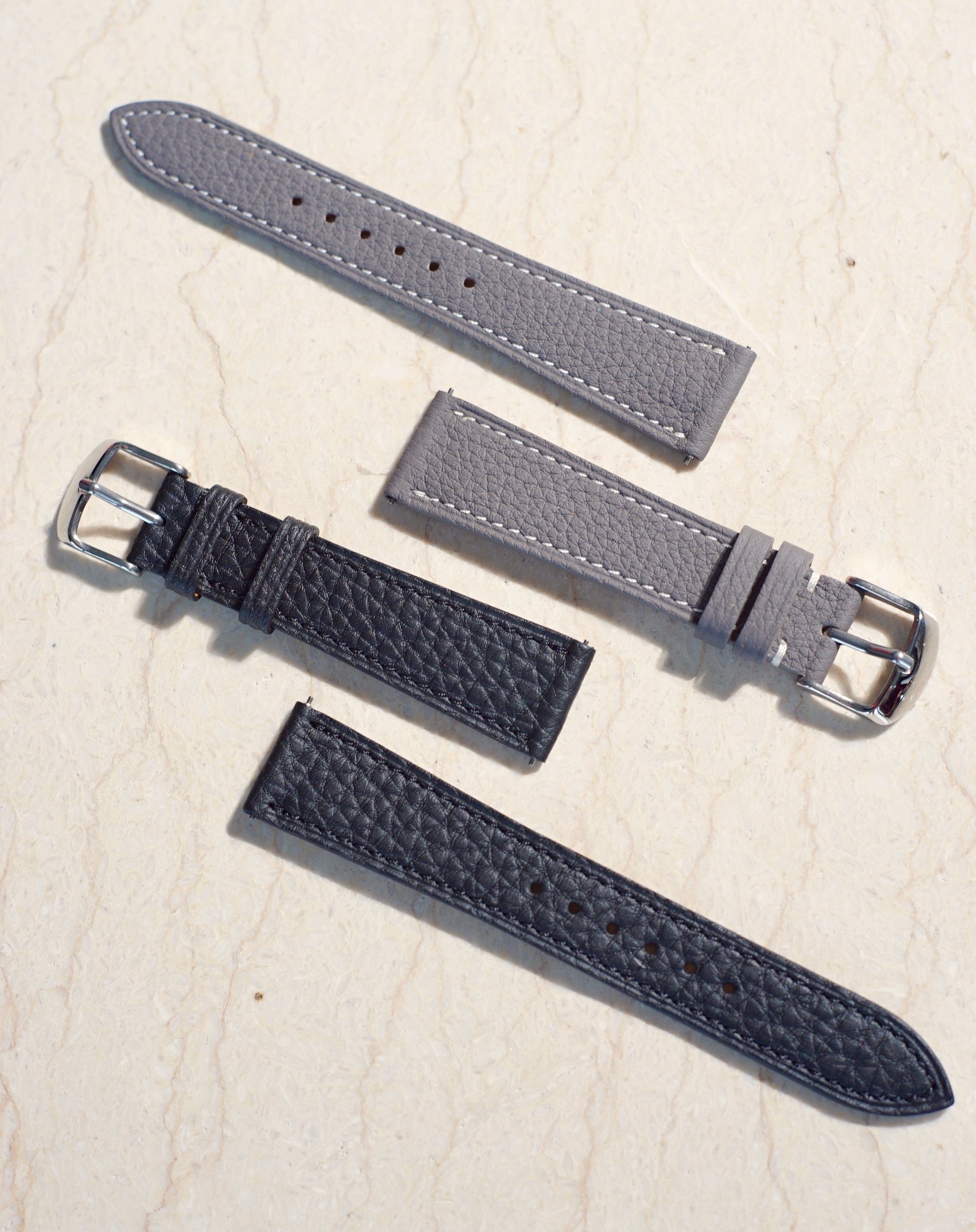 A pair of Black and Grey Quaint & Collectible Togo leather watch straps  laid flat on marble surface. Paired with silver polished buckle and quick release spring bar. 