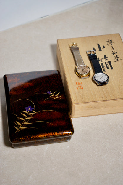 Two watches on a wooden box with decorative lid featuring floral designs.