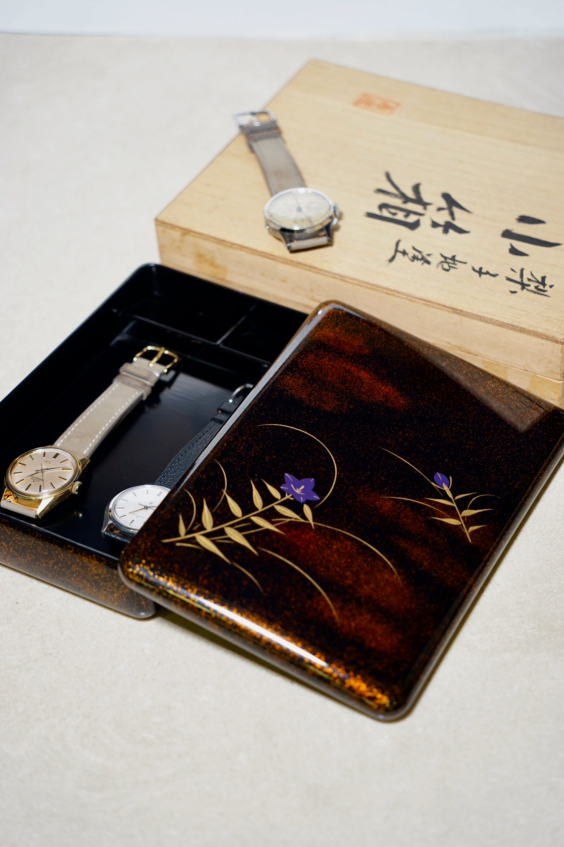 Two watches on a japanese urushi watch decorative wooden box with floral designs.