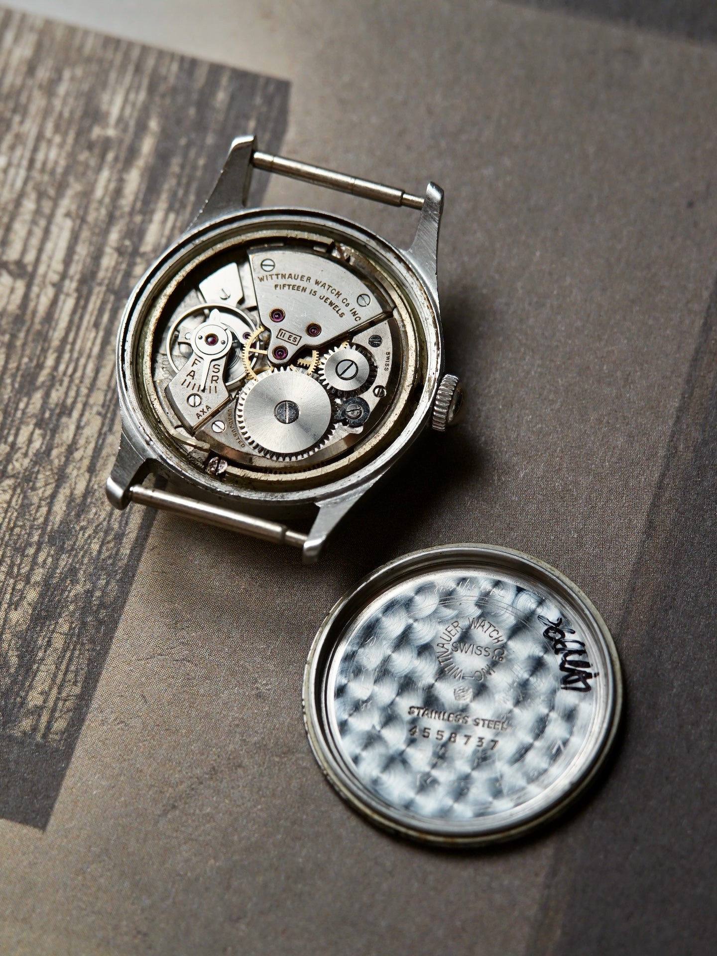 A vintage mechanical watch with its case removed, showing the exposed silver-toned movement and internal gears. The textured silver watch back is placed beside the open case on a brown paper surface.