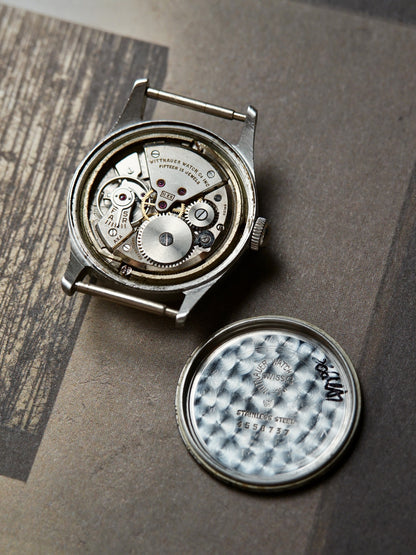 A vintage mechanical watch with its case removed, showing the exposed silver-toned movement and internal gears. The textured silver watch back is placed beside the open case on a brown paper surface.