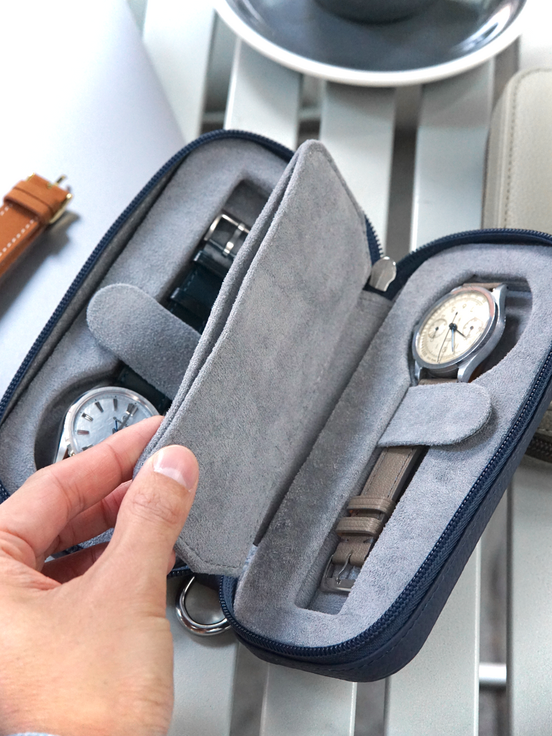 Gray watch case with a watch inside, held by a hand on a light-colored surface.